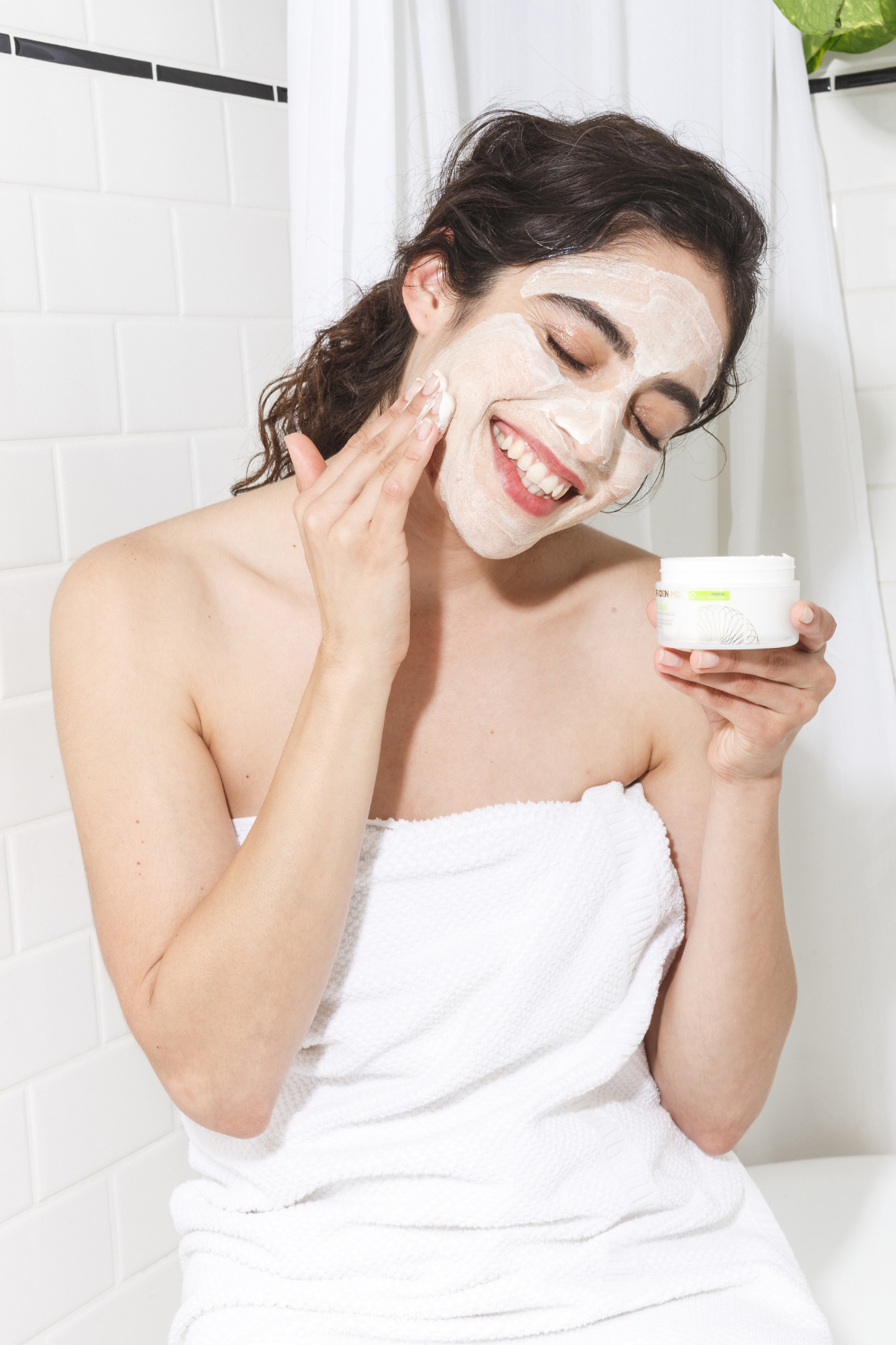 Woman applying cream to her face with a white towel wrapped around her shoulders.